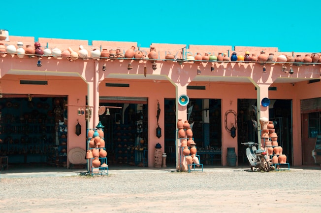 A market stall displays a variety of pottery items, including pots, vases, and decorative items arranged neatly on stands and shelves. The building features a pink facade with arches, and the atmosphere suggests it is a sunny day with a clear blue sky. Several pottery items are also lined up on the roof. A scooter is parked in front of the stall, and the ground appears dusty.