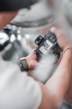 A person holding a digital camera with an image displayed on the screen. The camera is gripped in both hands and a blurred background creates a soft focus effect. The person wears a wristband and a tattoo is partially visible on the arm.