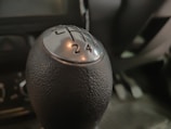 A close-up view of a car's manual gear shift knob, highlighting the numbers and gear positions in focus. The texture of the knob is visible, and the background shows a blurred interior of the vehicle.