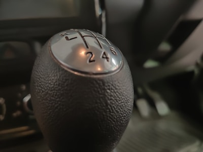 A close-up view of a car's manual gear shift knob, highlighting the numbers and gear positions in focus. The texture of the knob is visible, and the background shows a blurred interior of the vehicle.