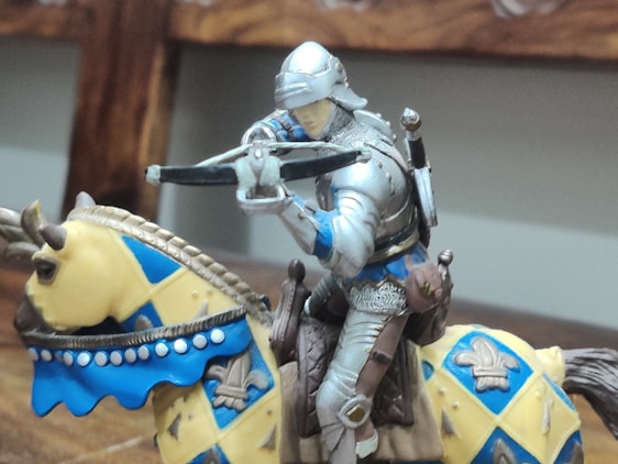 A medieval knight figurine in shiny armor is mounted on a horse. The knight is holding a crossbow, poised and ready to shoot. The horse is adorned with a vibrant blue and yellow decorative covering featuring a fleur-de-lis pattern. The setting appears indoors, likely on a wooden surface.