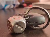 A metallic die keychain attached to a black car key lies on a dark surface, with a blurred background.