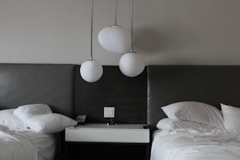 A modern bedroom setup featuring two beds with sleek, gray headboards. Between the beds, there is a nightstand with a digital clock, telephone, and a power outlet. Above, three round white pendant lights are suspended, adding a contemporary touch.