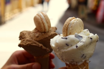 Two ice cream cones are held close together, each topped with a macaron. The cone on the left has a scoop of chocolate ice cream and a brown macaron, while the cone on the right features a scoop of vanilla ice cream with chocolate chips and a white macaron. The blurred background suggests an outdoor setting.