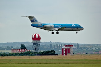 A KLM airplane is in mid-air, approaching for landing over a grassy field. In the background, there is an air traffic control tower with a red and white striped dome and additional buildings.