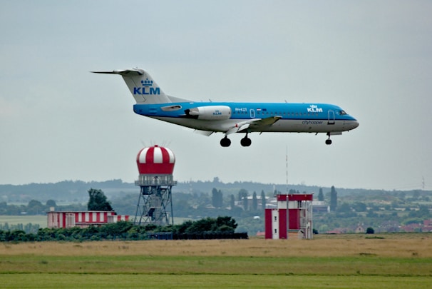 A KLM airplane is in mid-air, approaching for landing over a grassy field. In the background, there is an air traffic control tower with a red and white striped dome and additional buildings.