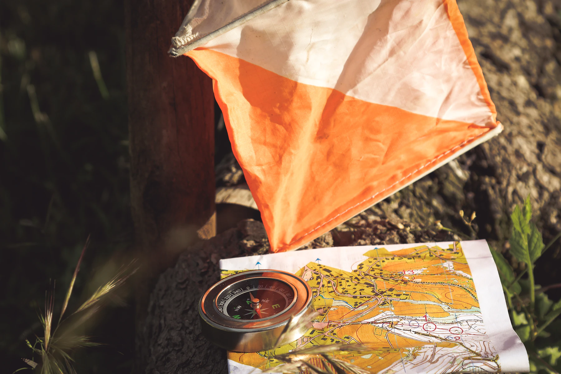 An orange-and-white checkpoint flag beside a compass and an orienteering map on a rock