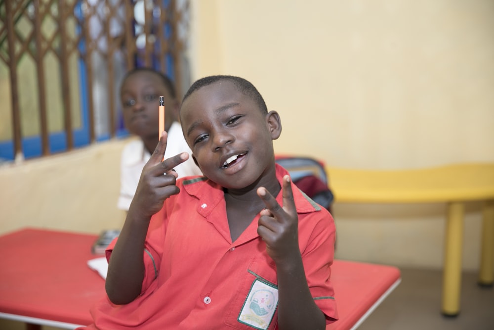 boy standing and making peace hand sign while holding pencil near another boy sitting inside room