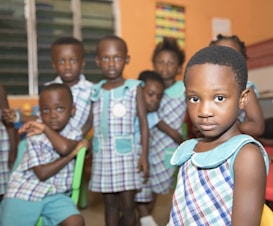 A group of young children in a classroom setting, wearing matching plaid uniforms with turquoise collars. The room has an orange-colored wall and windows with blinds. The children appear to be posing casually, displaying expressions ranging from serious to curious.