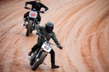 Two motorcyclists are engaged in a race on a dirt track. The rider in front, wearing a green jacket and black helmet, is leaning into a turn, maintaining balance with an extended foot. The second rider follows behind, also in a leaning position, showing concentration and control. Both bikes have number plates indicating their racing numbers.