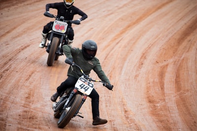 Two motorcyclists are engaged in a race on a dirt track. The rider in front, wearing a green jacket and black helmet, is leaning into a turn, maintaining balance with an extended foot. The second rider follows behind, also in a leaning position, showing concentration and control. Both bikes have number plates indicating their racing numbers.