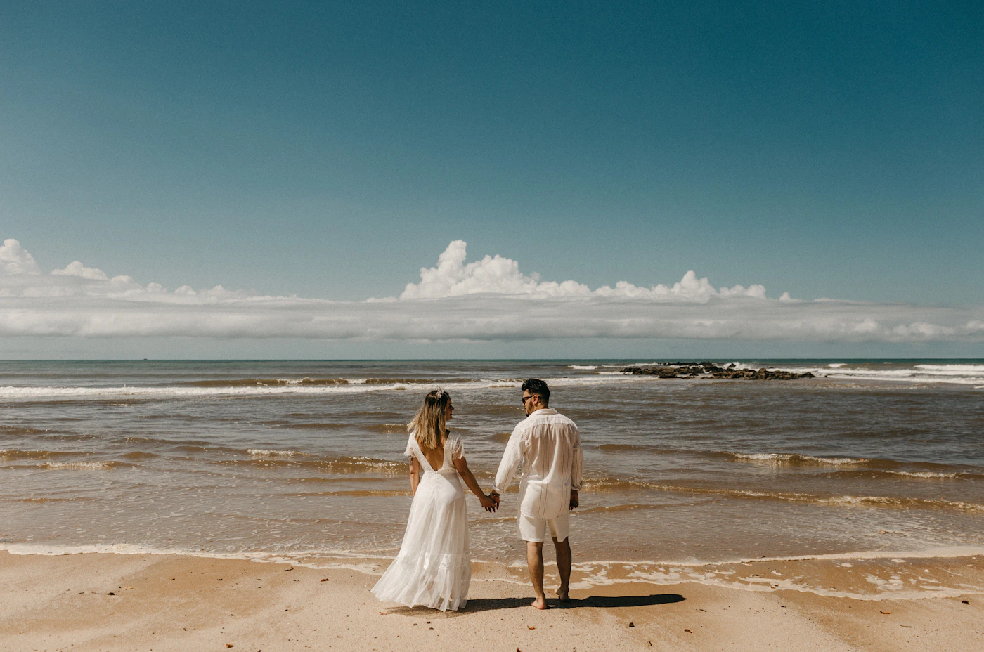 Couple in elopement attire on a Colombian Pacific beach in clear morning light with the jungle-covered hills visible along the coastline