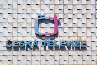 A textured wall composed of a grid of square tiles, with a prominent logo resembling a stylized television screen in a mix of purple and blue tones. Below the logo, the words 'ČESKÁ TELEVIZE' are displayed in blue capital letters.