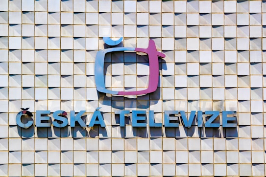 A textured wall composed of a grid of square tiles, with a prominent logo resembling a stylized television screen in a mix of purple and blue tones. Below the logo, the words 'ČESK&Aacute; TELEVIZE' are displayed in blue capital letters.
