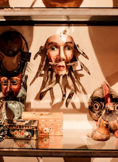 A collection of eclectic and colorful masks and decorative boxes are displayed on a glass shelf. The central mask has a human-like face with green eyes and tentacle-like appendages, surrounded by other masks and artisan-crafted items with diverse designs.