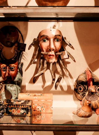 A collection of eclectic and colorful masks and decorative boxes are displayed on a glass shelf. The central mask has a human-like face with green eyes and tentacle-like appendages, surrounded by other masks and artisan-crafted items with diverse designs.