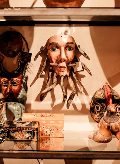 A collection of eclectic and colorful masks and decorative boxes are displayed on a glass shelf. The central mask has a human-like face with green eyes and tentacle-like appendages, surrounded by other masks and artisan-crafted items with diverse designs.