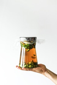 A hand holding a large glass jar filled with a liquid and sliced green fruits. The jar is topped with a metal lid and covered with plastic wrap.