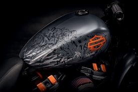 A motorcycle fuel tank with an intricate design featuring graffiti-style art in black and gray tones along with a prominent orange logo. The tank is attached to a dark-colored motorcycle with parts of the seat and engine visible.