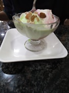 A glass dessert cup filled with colorful ice cream scoops, topped with almonds, placed on a white square plate. The table surface is dark and glossy.