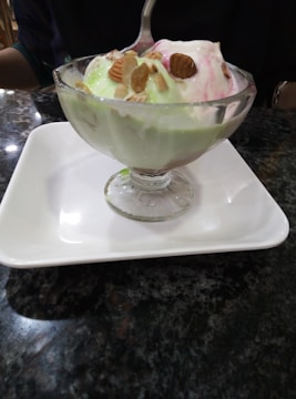 A glass dessert cup filled with colorful ice cream scoops, topped with almonds, placed on a white square plate. The table surface is dark and glossy.
