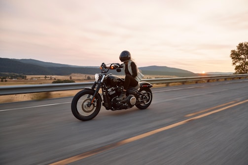 A lone motorcyclist rides along an open highway with a backdrop of expansive fields and distant hills under a soft sunset. The rider wears a helmet and protective gear, suggesting a focus on safety and adventure. The road is empty, emphasizing the tranquility and freedom of the journey.