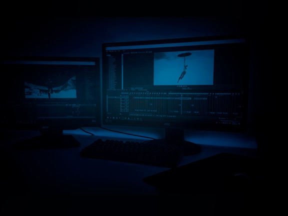 A dimly lit room featuring two computer monitors displaying video editing software. The screens show footage and timelines, with one monitor depicting an image of a person with an umbrella. A keyboard is visible in front of the monitors.