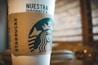A close-up of a coffee cup featuring the Starbucks logo and a cardboard sleeve. The design includes the text 'NUESTRA PROMESA' in prominent letters. The background is softly blurred, with warm earth tones creating a cozy, inviting atmosphere.