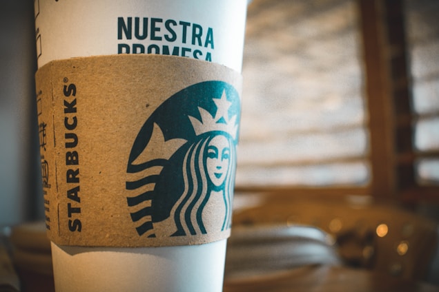 A close-up of a coffee cup featuring the Starbucks logo and a cardboard sleeve. The design includes the text 'NUESTRA PROMESA' in prominent letters. The background is softly blurred, with warm earth tones creating a cozy, inviting atmosphere.