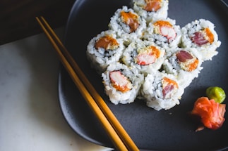 A plate featuring sushi rolls, delicately arranged with slices of raw fish, cream cheese, and rice, garnished with wasabi and pickled ginger. Accompanied by wooden chopsticks resting on the edge of a dark ceramic plate.