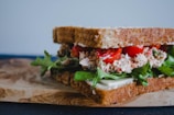 A close-up of a sandwich with layers of brown bread, containing fresh lettuce leaves, slices of red tomatoes, and a creamy filling. The sandwich is placed on a wooden board.