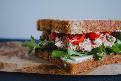 A close-up of a sandwich with layers of brown bread, containing fresh lettuce leaves, slices of red tomatoes, and a creamy filling. The sandwich is placed on a wooden board.
