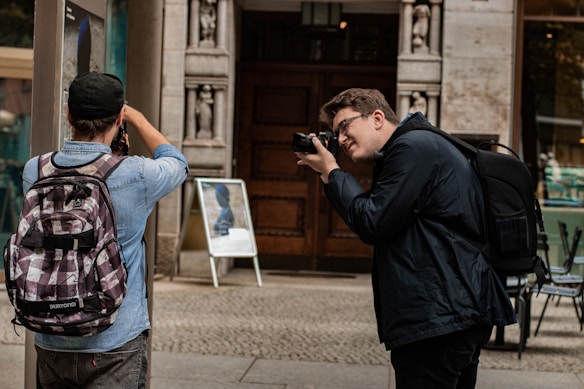 Two people, each holding a camera, are engaged in taking photographs on a city street. One person is wearing a denim shirt and a patterned backpack, while the other is in a dark jacket with a black backpack. They stand near a building with stone carvings beside a sidewalk. An A-frame sign displaying a blurred image is visible in the background.