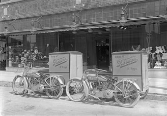 Two vintage Harley-Davidson motorcycles are parked in front of a storefront with large display windows. The motorcycles have attached delivery boxes labeled 'Shop At Parisian.' Inside the window display, various hats are arranged, suggesting a hat shop.