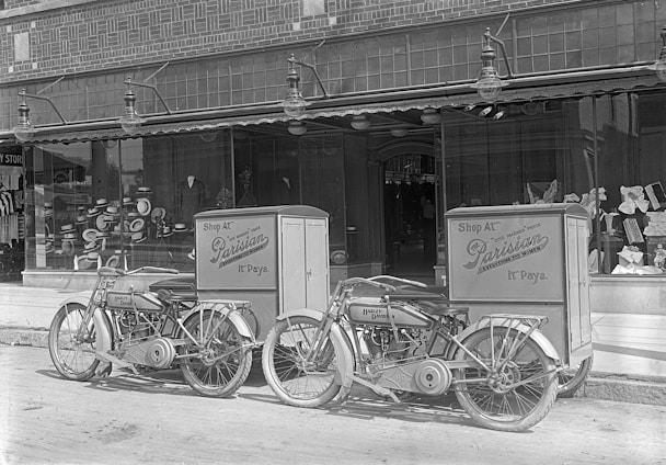 Two vintage Harley-Davidson motorcycles are parked in front of a storefront with large display windows. The motorcycles have attached delivery boxes labeled 'Shop At Parisian.' Inside the window display, various hats are arranged, suggesting a hat shop.
