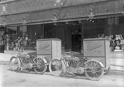 Two vintage Harley-Davidson motorcycles are parked in front of a storefront with large display windows. The motorcycles have attached delivery boxes labeled 'Shop At Parisian.' Inside the window display, various hats are arranged, suggesting a hat shop.