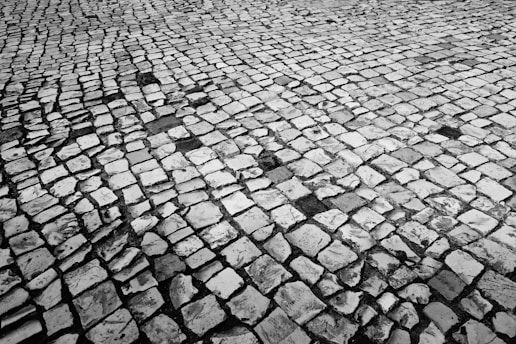 A close-up view of a cobblestone pathway featuring irregularly shaped stones arranged in a repetitive pattern. The stones are weathered and show signs of wear, with varying shades of gray creating a textured visual effect.