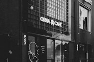 A black and white photograph of a café with a modern and minimalist exterior. The café's name is displayed prominently in bright, bold letters above the entrance. The building has a grid-like pattern on its exterior walls, giving it a contemporary feel. Large glass windows reveal the interior, and there is a decorative abstract design beside the entrance.