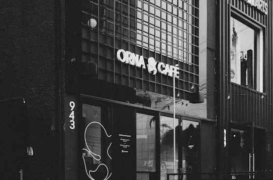 A black and white photograph of a caf&eacute; with a modern and minimalist exterior. The caf&eacute;'s name is displayed prominently in bright, bold letters above the entrance. The building has a grid-like pattern on its exterior walls, giving it a contemporary feel. Large glass windows reveal the interior, and there is a decorative abstract design beside the entrance.