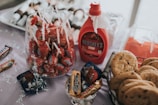 A table is set with an assortment of sweet treats, including lollipops in a glass container, a bottle of Hershey's syrup, wrapped candies, and a basket of chocolate chip cookies. There is also a tray of skewered snacks in the background.