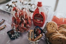 A table is set with an assortment of sweet treats, including lollipops in a glass container, a bottle of Hershey's syrup, wrapped candies, and a basket of chocolate chip cookies. There is also a tray of skewered snacks in the background.