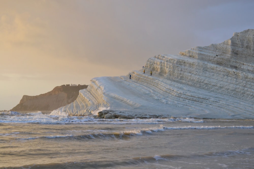 poucas pessoas caminhando na montanha coberta de neve perto do mar sob céus cinzas e laranjas