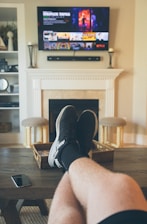 A person is relaxing in a living room with feet up on a wooden table. A TV mounted above a fireplace is displaying a popular streaming platform. Shelves on either side of the fireplace hold decorative items. A smartphone can be seen on the table.