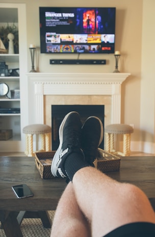 A person is relaxing in a living room with feet up on a wooden table. A TV mounted above a fireplace is displaying a popular streaming platform. Shelves on either side of the fireplace hold decorative items. A smartphone can be seen on the table.
