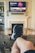 A person is relaxing in a living room with feet up on a wooden table. A TV mounted above a fireplace is displaying a popular streaming platform. Shelves on either side of the fireplace hold decorative items. A smartphone can be seen on the table.