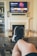 A person is relaxing in a living room with feet up on a wooden table. A TV mounted above a fireplace is displaying a popular streaming platform. Shelves on either side of the fireplace hold decorative items. A smartphone can be seen on the table.