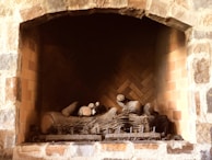 A stone fireplace with a neatly arranged set of logs inside. The interior is lined with brickwork in a herringbone pattern, providing a rustic appearance. The warm tones of the bricks and stones add to the cozy atmosphere.