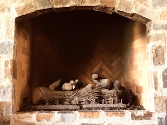 A stone fireplace with a neatly arranged set of logs inside. The interior is lined with brickwork in a herringbone pattern, providing a rustic appearance. The warm tones of the bricks and stones add to the cozy atmosphere.