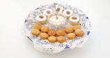 A decorative plate with white lace holds an assortment of mini cupcakes with white frosting and brown decorative swirls, alongside small round pastries arranged neatly. A small bowl of white creamy dip with a spoon is placed in the center.