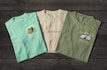 Three t-shirts are displayed on a dark wooden surface. The t-shirts have different prints, with the first one being light turquoise with the word 'Doheny', an orange fish graphic, and the text 'The Lagoon'. The second shirt is off-white featuring 'Starboard Creek' across the chest. The third t-shirt is olive green with the words 'La Jolla' and an image of a kayak with 'The Cove' written underneath.